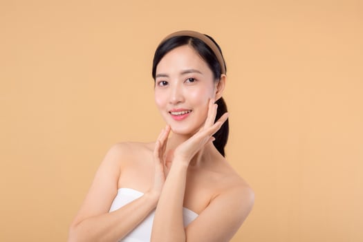 Beautiful girl asian model touching fresh glowing hydrated facial skin on beige background closeup. Beauty face young woman with natural makeup and healthy skin portrait. Skin care concept photo