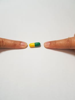 Isolated white photo of two forefingers pointing at a green and yellow medicine capsule.