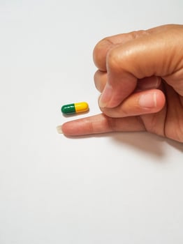 Isolated white photo of a little finger measuring the size of a green and yellow medicine capsule.