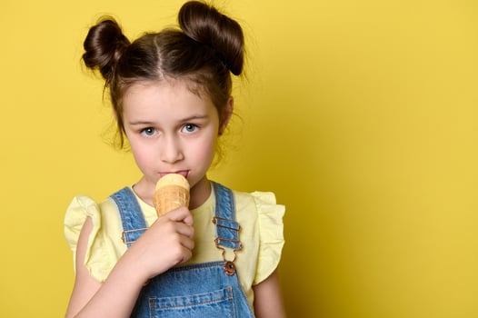 Close-up little child girl licking scoop of ice cream in waffle cone, looking at camera, isolated over yellow background photo