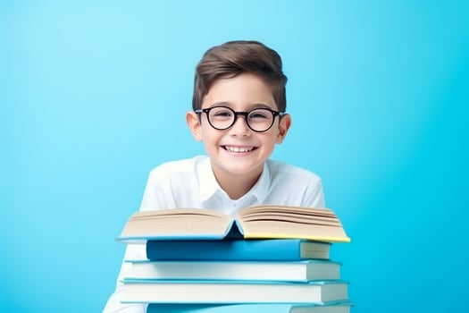portrait of a happy child little boy with glasses sitting on a stack of books and reading a books, light blue background. photo