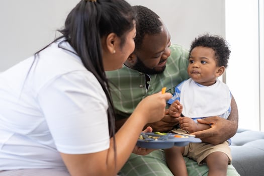 Asian mother feeding her 9 months old her cute little baby and African American helping for holding food plate At Home. Photo series of family, kids and happy people concept. Parents feed kids.