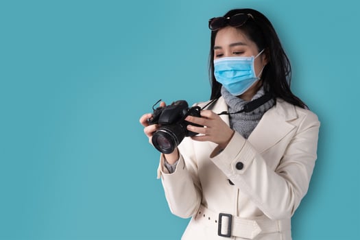 Young beautiful Asian woman holding a camera and wearing a medical face mask on a blue isolated background, Concept of traveler tourist and vacation. photo