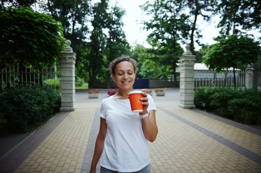 Smiling beautiful confident woman tourist walking in the park alley, holding orange cardboard mug with takeaway drink photo