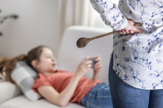 Adolescent girl ignores her mother standing upset with a wooden spoon next to her and keeps being on her mobile phone photo