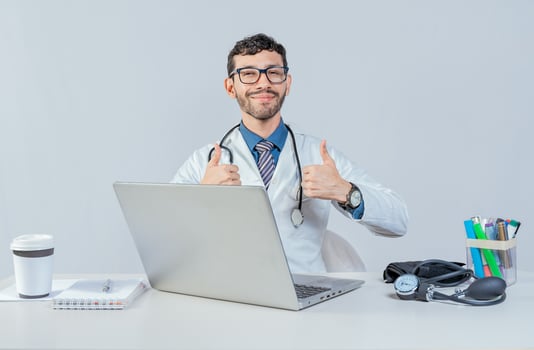 Smiling doctor sitting with laptop giving thumbs up. Male doctor with laptop showing thumbs up in ok gesture. photo
