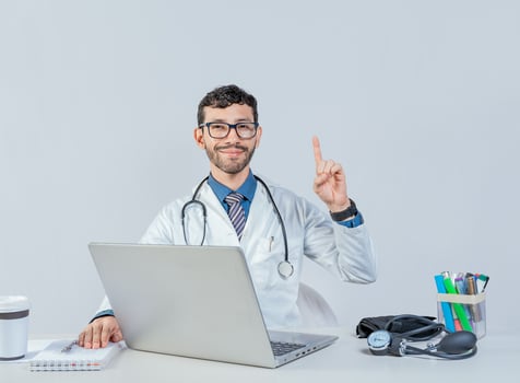 Smiling doctor with laptop with finger up in idea gesture. Doctor sitting with laptop pointing up isolated. Young doctor sitting with laptop with an idea gesture. photo