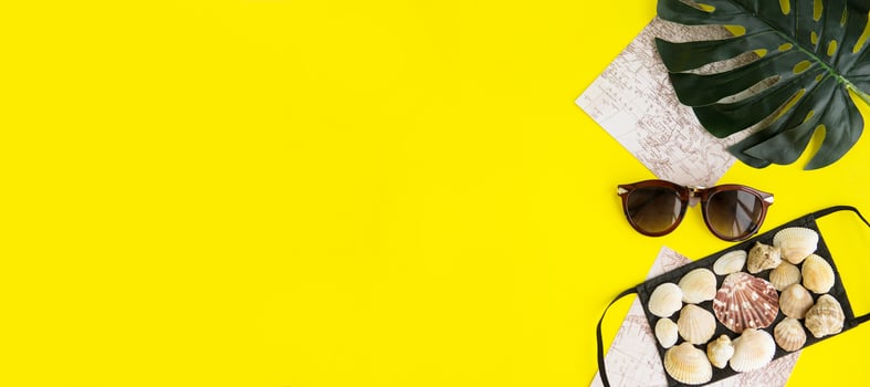 Black medical face mask and seashells, sunglasses, monstera leaf on a yellow background. Flat lying, copy space, vacation cancellation, pandemic, tourism. photo