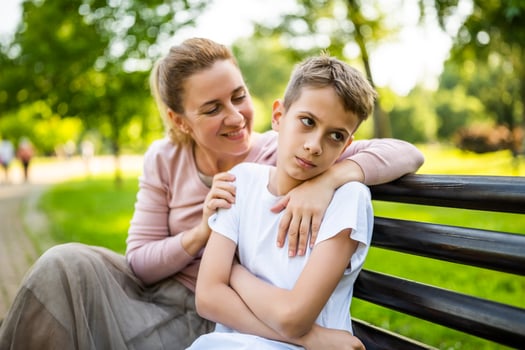 Happy mother is sitting with her son on bench in park. Boy is offended and pouting. photo