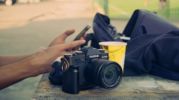 Fujifilm X T30 camera placed on a wooden surface with a bag in the background and a hand holding a smartphone. photo