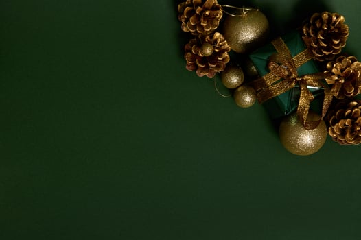 Flat lay of a present in shiny wrapping gift paper with golden bow, pines cones and gold Christmas tree toys laid out on the corner of green background. Copy space for advertisement photo