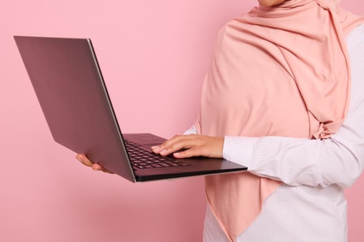 Close-up portrait of a woman wearing pink hijab standing against colored background with an open laptop in hands and typing text.Concept of successful Muslim programmer woman, worker at IT technology photo