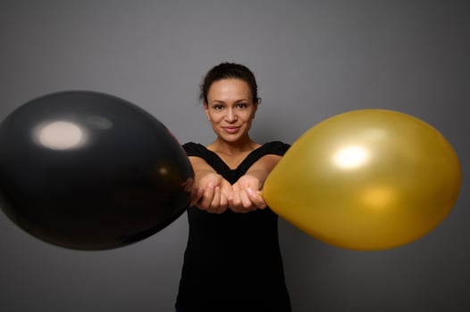 Beautiful woman dressed in black smiles looking at camera and poses against gray background holding gold and black air balloons in her outstretched hand. Black Friday concept with copy ad space photo