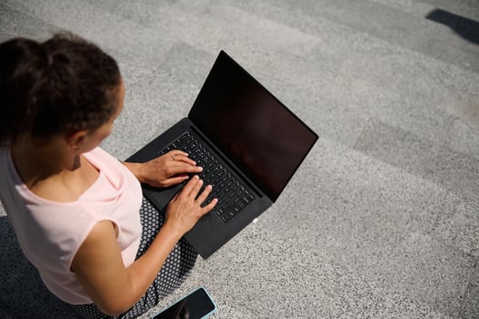 Top view of a woman sitting on steps and typing text on on laptop with blank black monitor screen with copy space for advertising on the urban background. Business concept photo