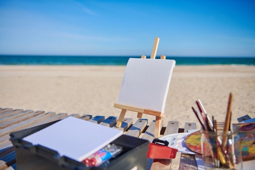 Blank white canvas on a wooden table easel next to paints and brushes on a wooden deck chair on the beach against the background of beautiful blue sea photo