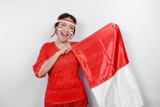 Happy smiling Indonesian woman wearing red kebaya and headband holding Indonesia's flag to celebrate Indonesia Independence Day isolated over white background. photo