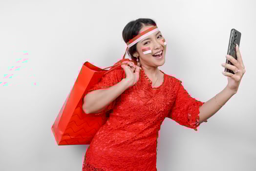An excited Asian woman wearing red kebaya holding a shopping bag and her smartphone, studio shot isolated on white background. Indonesia's Independence day concept photo