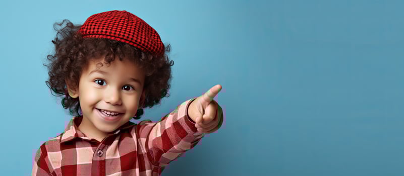Happy young boy with curly hair pointing up wearing a red checkered shirt isolated on a blue background in a studio portrait photo
