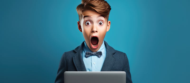 A surprised teenage boy holds an open laptop suggesting an educational concept with a blue studio background photo