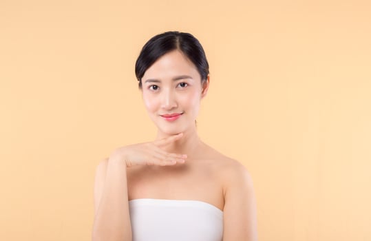 Beautiful girl asian model touching fresh glowing hydrated facial skin on beige background closeup. Beauty face young woman with natural makeup and healthy skin portrait. Skin care concept photo