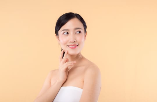 Beautiful girl asian model touching fresh glowing hydrated facial skin on beige background closeup. Beauty face young woman with natural makeup and healthy skin portrait. Skin care concept photo