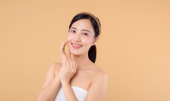 Beautiful girl asian model touching fresh glowing hydrated facial skin on beige background closeup. Beauty face young woman with natural makeup and healthy skin portrait. Skin care concept photo