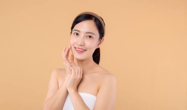 Beautiful girl asian model touching fresh glowing hydrated facial skin on beige background closeup. Beauty face young woman with natural makeup and healthy skin portrait. Skin care concept photo