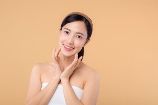 Beautiful girl asian model touching fresh glowing hydrated facial skin on beige background closeup. Beauty face young woman with natural makeup and healthy skin portrait. Skin care concept photo