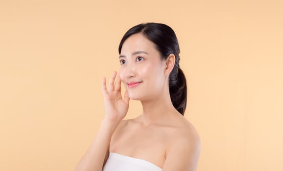 Beautiful girl asian model touching fresh glowing hydrated facial skin on beige background closeup. Beauty face young woman with natural makeup and healthy skin portrait. Skin care concept photo