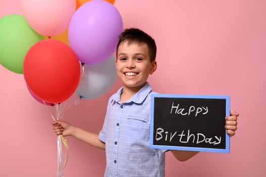 Happy teenage boy holds multicolored colorful balloons in one hand and a chalkboard with lettering Happy Birthday in the other. Isolated over pink background with copy space photo