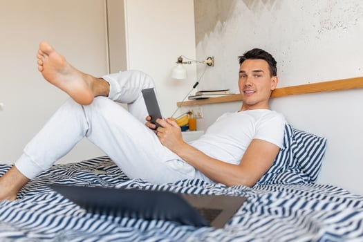 attractive young smiling man in casual pajamas outfit sitting in bed in morning working on laptop photo