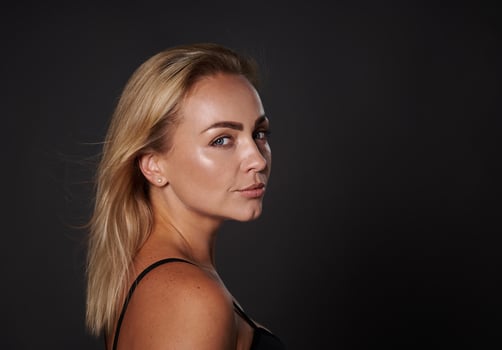 Close-up of confident blonde beautiful middle aged woman with healthy tanned clean glowing skin in black bra looking at camera standing three quarters on dark background with copy space. Studio shot photo
