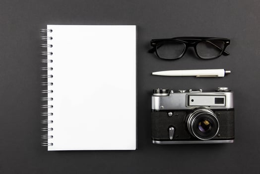 Modern office workplace flat lay with blank spiral paper notebook mockup, retro camera, pen and reading glasses on dark black background. Top view. Copy space. photo