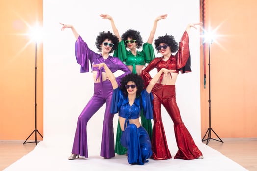 A group of girls in colorful flared suits and afro wigs pose in a photo studio. Disco style from the eighties or seventies.