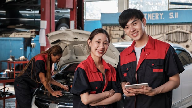 Two Asian professional mechanics, male engineer and partner smile and look at camera, inspect repair work checklist at garage, service car maintenance, and fixing specialist occupations auto industry. photo