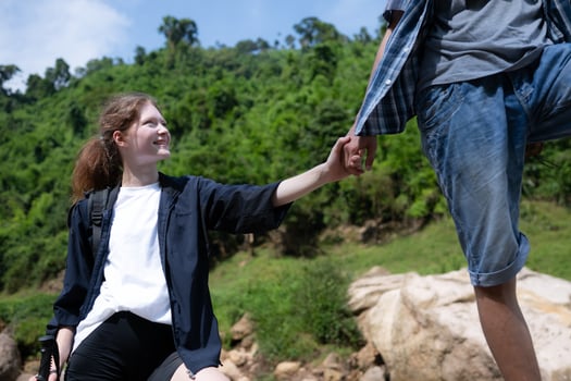 Young woman hiker with backpack and trekking poles with a young man helping to pull him up to stand on the rock photo