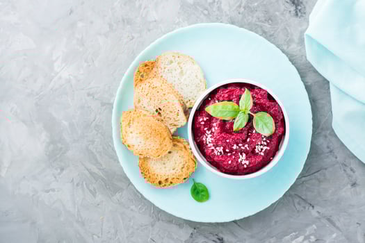 Homemade baked beetroot hummus in a bowl with sesame seeds and basil and baked small toast on a plate on the table. Top view photo