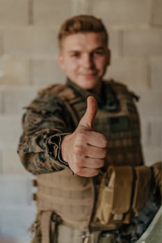 The soldier makes a gesture of success with his hand. A soldier in full war gear stands in front of a stone wall and shows the ok sign with his finger photo