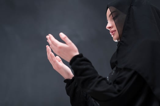 Portrait of beautiful muslim woman in fashionable dress with hijab making traditional prayer to God keeps hands in praying gesture isolated on black background photo