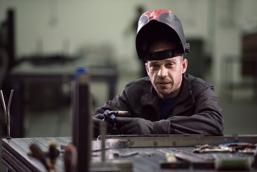 Portrait of Young Professional Heavy Industry Engineer. Worker Wearing Safety Vest and Hardhat Smiling on Camera. In the Background Unfocused Large Industrial Factory photo