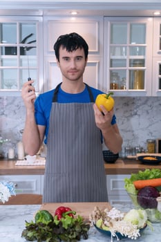 Happy young man wearing aprons at home cooking meal his hands holding kitchen knife and yellow sweet peppers look at camera. Lifestyle concept photo