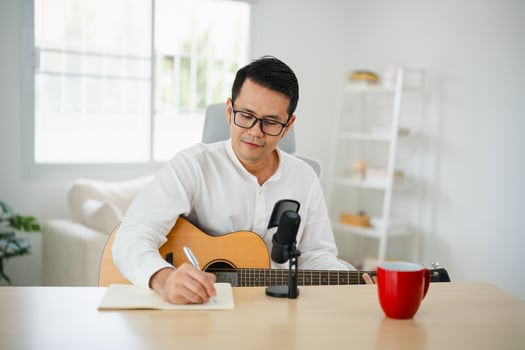Asian man wearing headphone play guitar and sing a song and learning online with laptop and notesbook and compose writing song, record sound and use computer recording music program. Musician concept. photo
