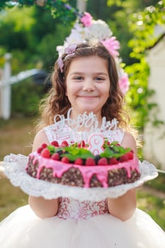 girl holding birthday cake with number 8 and inscription Happy Birthday. Celebrating ceremonial events on street during quarantine. child in background decorated photo zone of flowers.