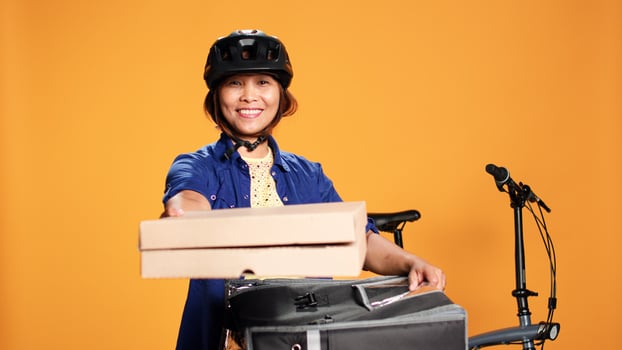 Friendly pizza delivery woman wearing bike gear bringing order to customer. Upbeat asian courier getting lunch boxes out of thermal food backpack, isolated over orange studio background photo