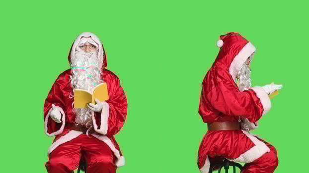 Saint nick reads lecutre novel on chair, man dressed as santa claus with red suit and glasses while he is reading literature book over greenscreen. Person in costume enjoying fairytale fiction. photo