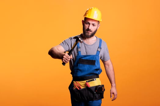 Construction worker using hammer on renovation project, standing over yellow background. Professional male renovator doing reconstruction work with sledgehammer and tools, studio shot. photo