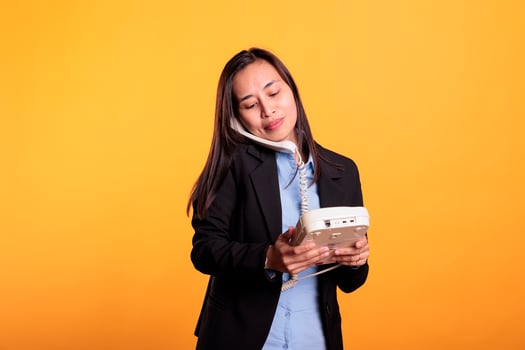 Asian woman in formal suit talking on landline phone call, using telephone with cord standing over yellow background. Confident young adult using office retro phone having remote discussion in studio photo