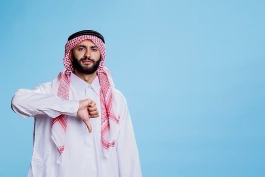 Muslim man dressed in traditional clothes standing with confident expression and thumb down portrait. Serious arab posing with gesture indicating disagreement and looking at camera photo