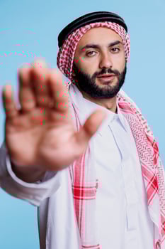 Confident muslim man making forbidden stop sign with palm and looking at camera. Serious arab person dressed in traditional clothes showing rejection gesture with hand studio portrait photo