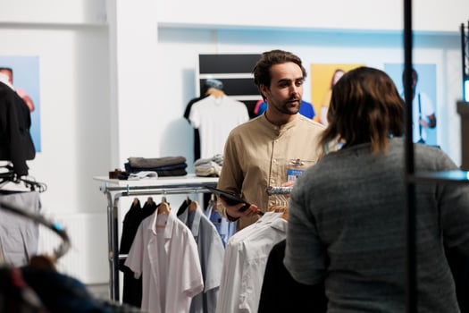 Mall worker standing near clothes rack and checking apparel available for purchase using digital tablet. Shopping center young man employee checking store app on portable gadget photo